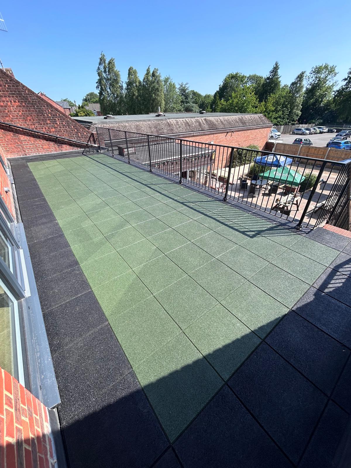Roof terrace surfaced with green rubber safety tiles bordered by black tiles and enclosed by metal railings