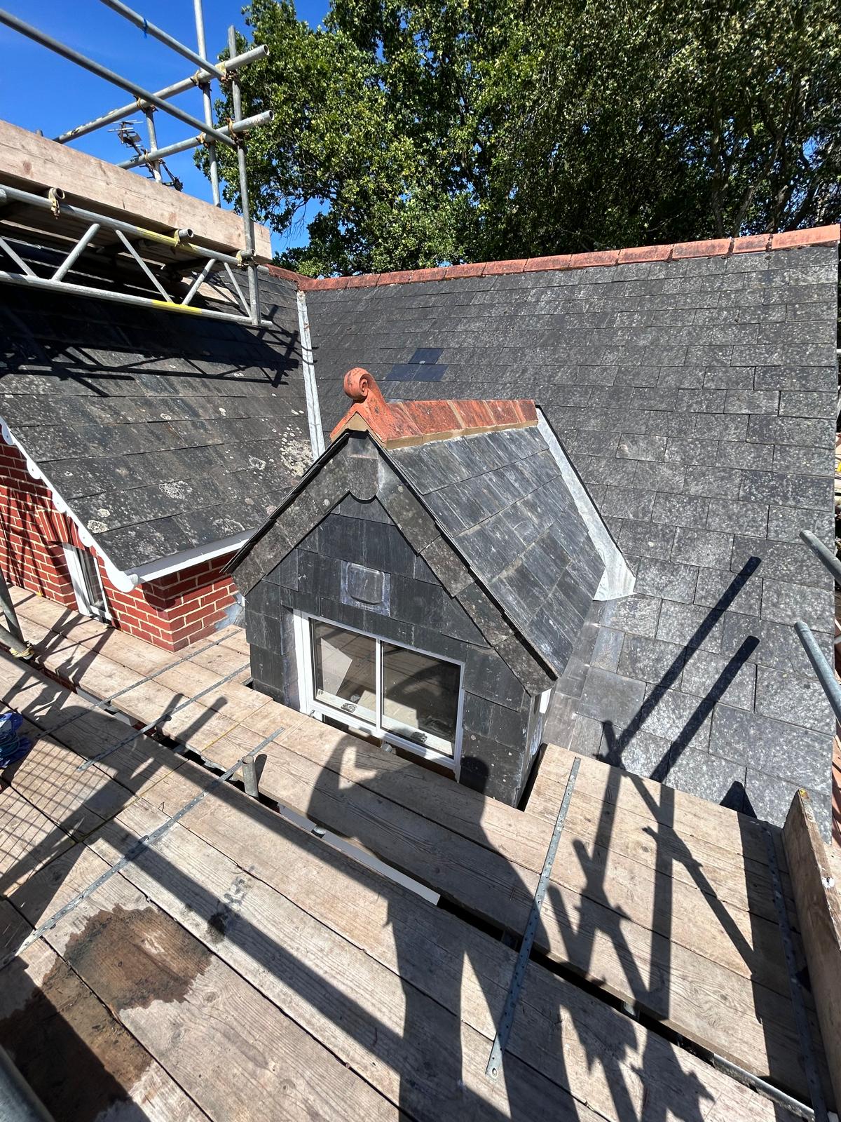 Slate-hung pitched dormer with a terracotta ridge and scroll finial, lead cheeks and soakers against the surrounding slate roof